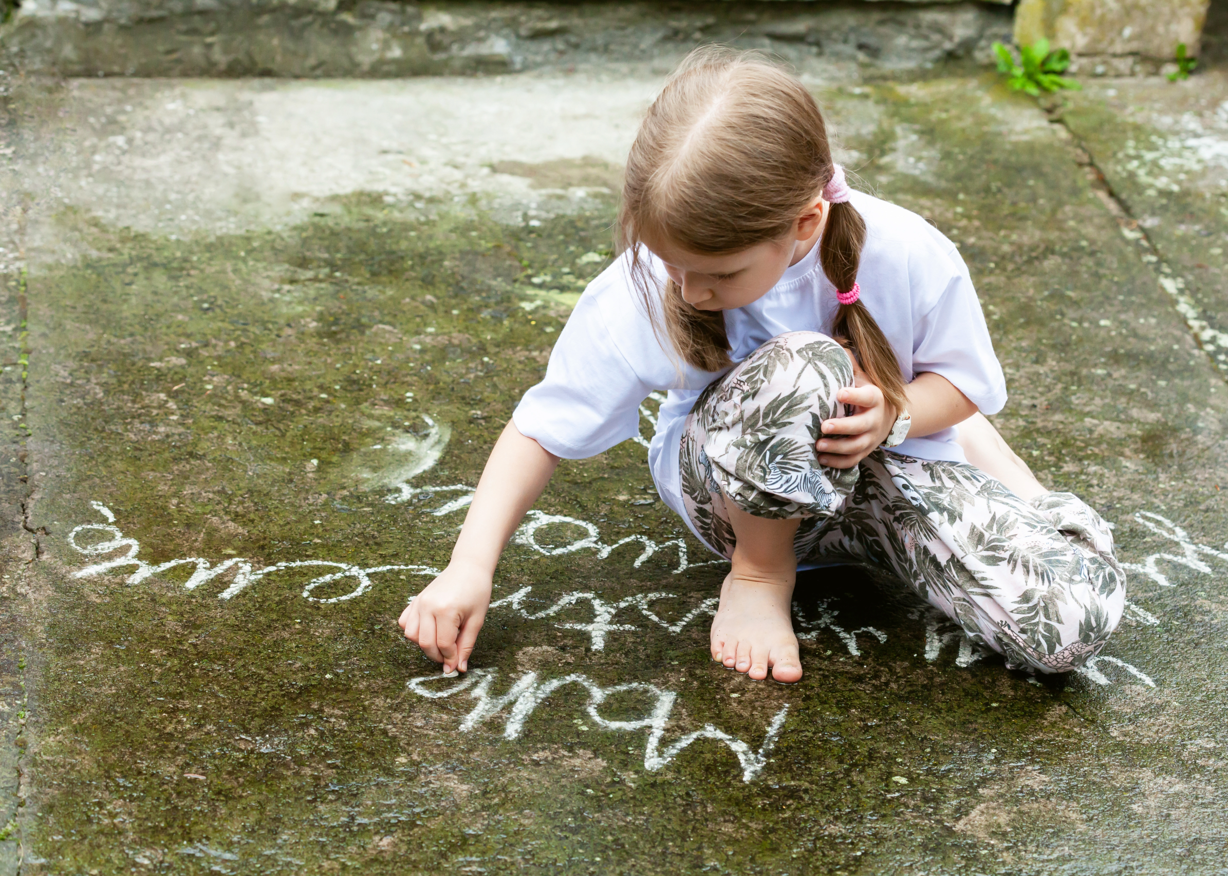 A child writing in chalk on a concrete surface outside in the sunshine.